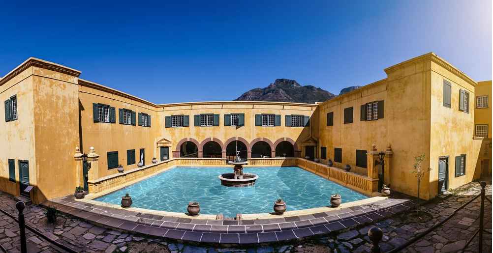 Inner courtyard of the Castle of Good Hope, Cape Town, showing the central Dolphin Pool fountain with dolphin motifs, surrounding yellow-painted walls, the dividing Kat wall, De Kat Balcony, and the area marked as Het Backhuys (bakery) on historical layouts